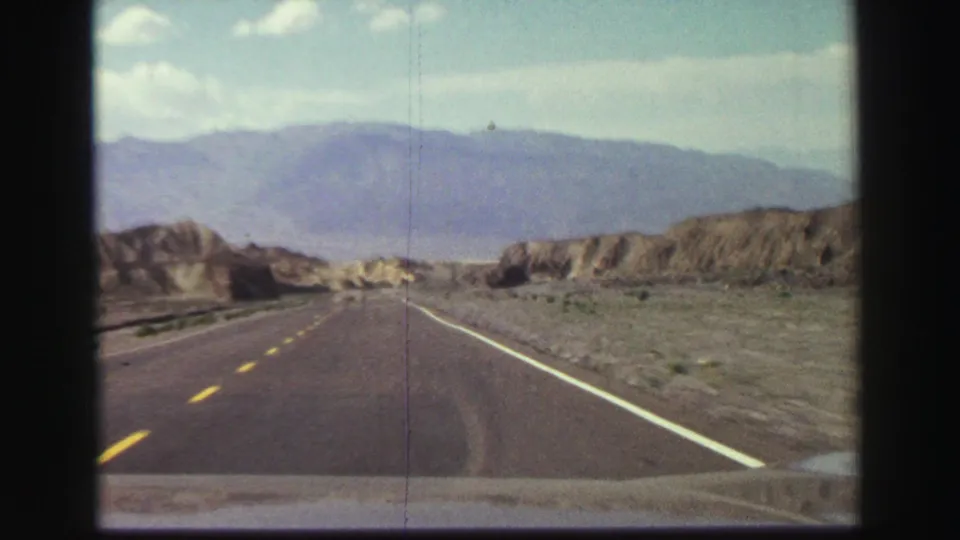Moving-car view of a paved two-lane road crossing arid foothills and distant mountains in the supplied 1969 California travel footage.