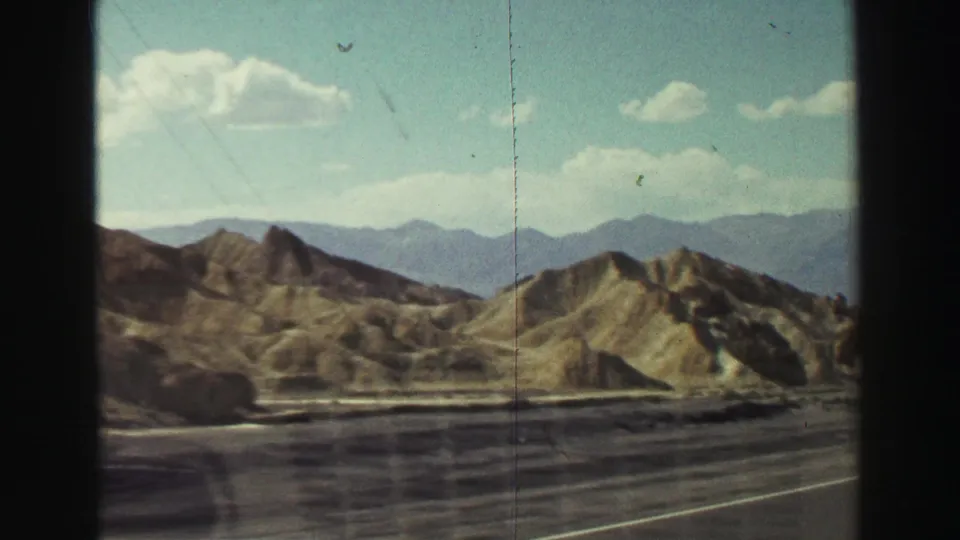 8mm roadside view of arid hills, a paved shoulder, and distant mountains in the supplied 1969 California home-movie footage.