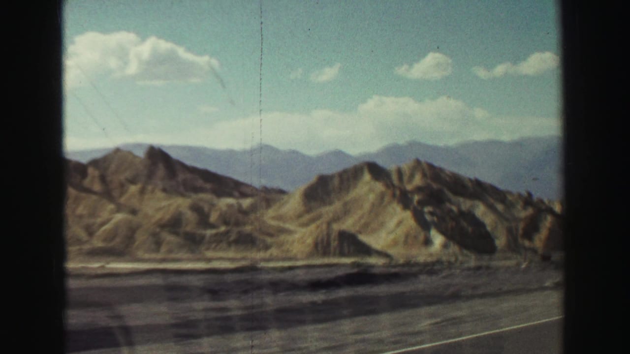 Rugged dry hills and distant mountains seen from a roadway under a blue sky.