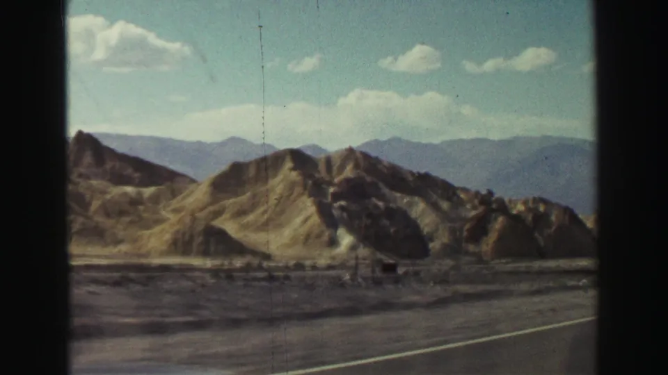 Moving-vehicle 8mm view of rugged hills and mountains near Lake Tahoe, California, with highway pavement crossing the lower frame.