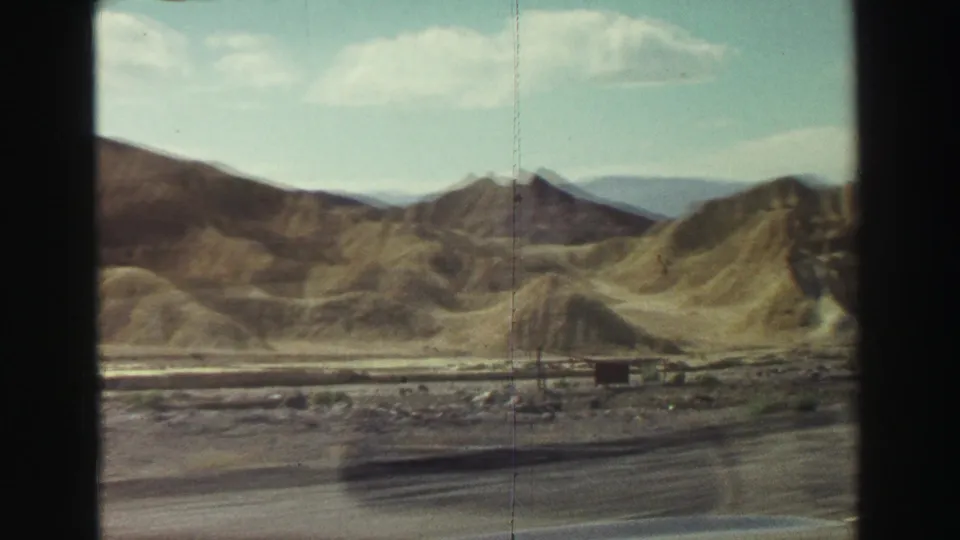 1969 home-movie frame from a moving car showing rolling dry hills, a roadside area, and a small distant structure or sign near Lake Tahoe, California.