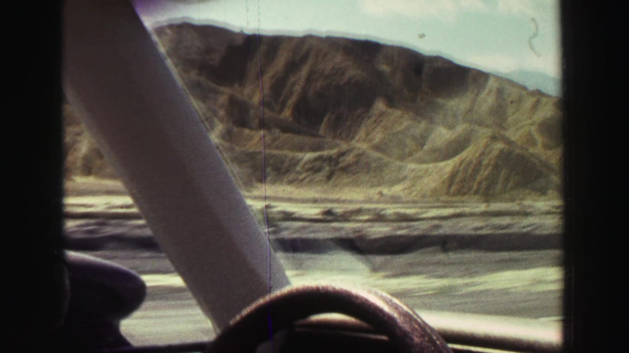 View through a car windshield toward dry ridged hills beside a road.