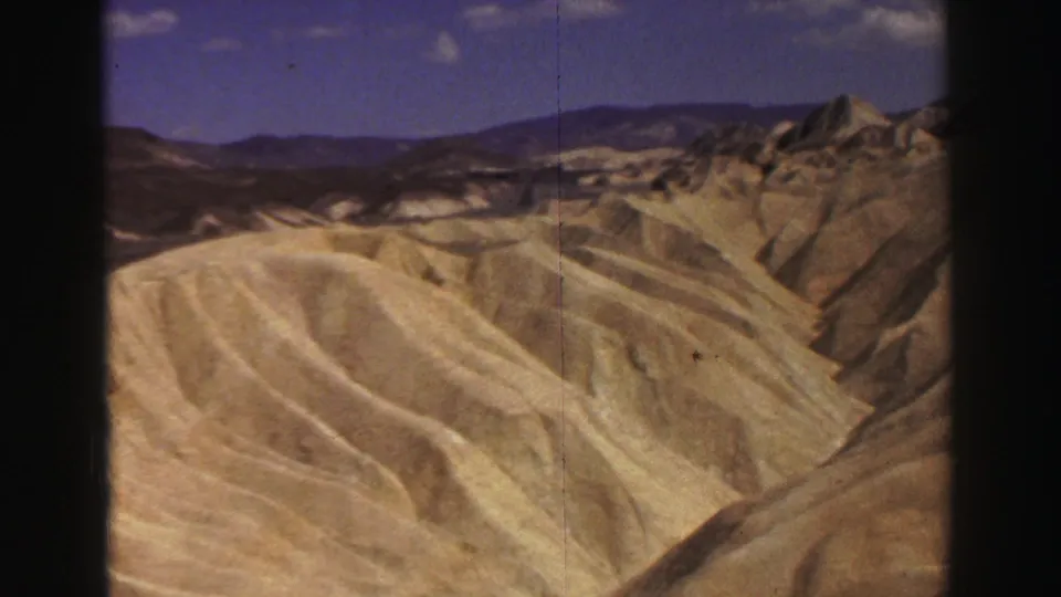 Arid California hillside landscape from 1969 home-movie footage, showing pale ridges, rocky formations and distant mountains.