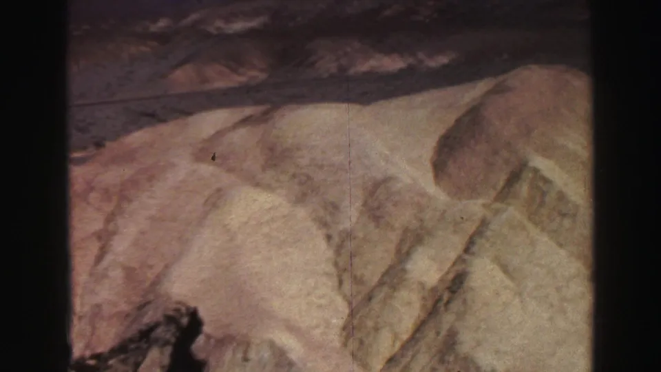 1969 8mm frame of eroded sandy terrain with pink and ochre tones, dry gullies, and a darker ridge line in the background.