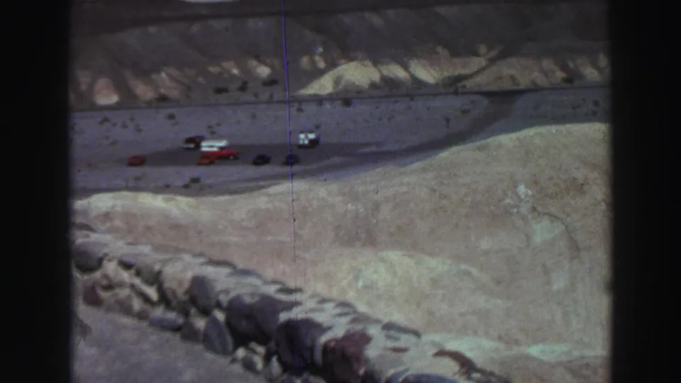 Visitor-shot 8mm frame of cars at a Lake Tahoe overlook road, with sandy terrain and retaining stones in view.