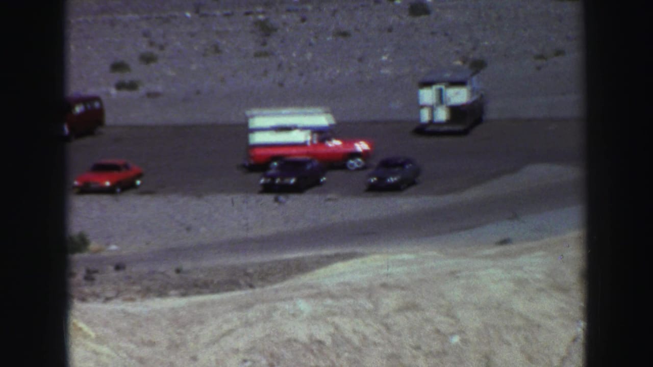 1969 Lake Tahoe roadside view with a red camper pickup, red sedan, and dark cars on gravel near pale mountain terrain.