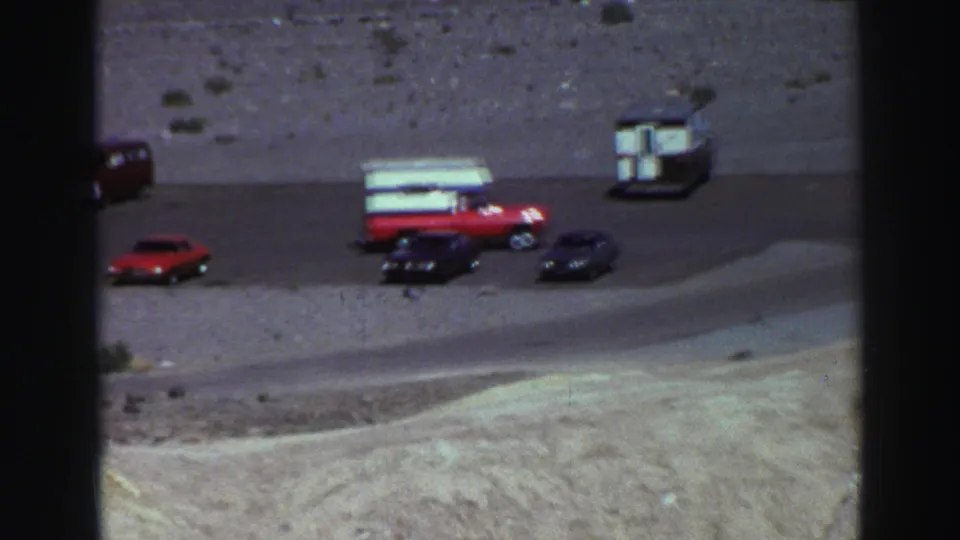 1969 Lake Tahoe roadside view with a red camper pickup, red sedan, and dark cars on gravel near pale mountain terrain.