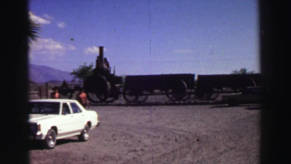 1969 Lake Tahoe-area home-movie frame showing visitors near a displayed steam locomotive, with a white sedan and mountain backdrop.