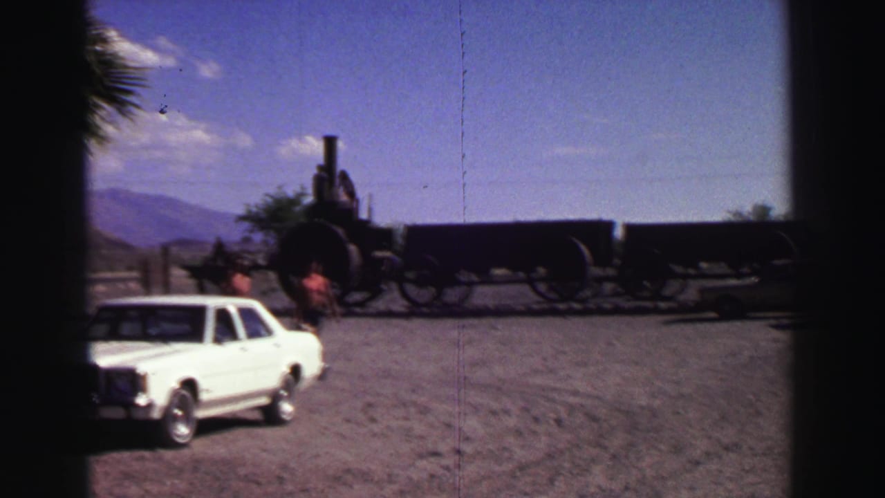 White sedan on a gravel lot near two people and a steam locomotive.