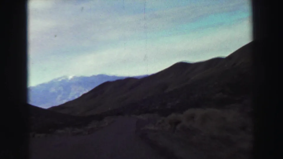 Vehicle-shot 1969 travel view of a paved road through dry hills with distant mountains and a bright sky.