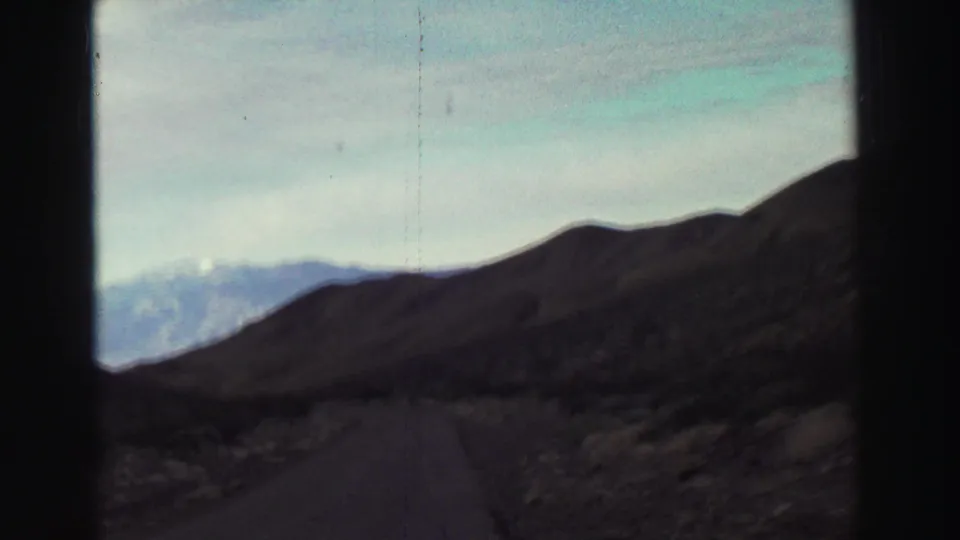 1969 home-movie road view showing a narrow paved route through dry hills with distant mountain ridges under pale sky.