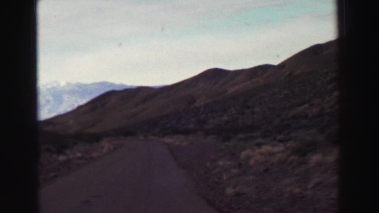 Paved mountain road seen through dark frame edges with dry hills and distant pale peaks.