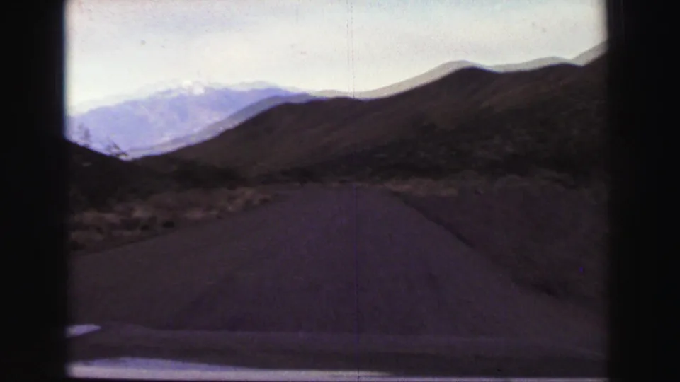 Moving-car 8mm frame near Lake Tahoe with a curving paved road, scrub-covered slopes and snow-streaked mountains in the distance.