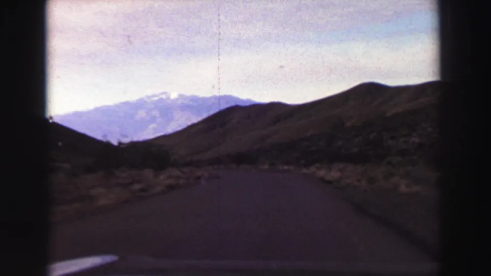 1969 home-movie road view near Lake Tahoe, with a paved lane, dry brush, dark hillside and snow-capped mountains beyond.