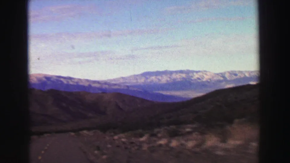 8mm Lake Tahoe-area vista from a moving car, showing a road through rounded hills and a distant snow-streaked mountain ridge.
