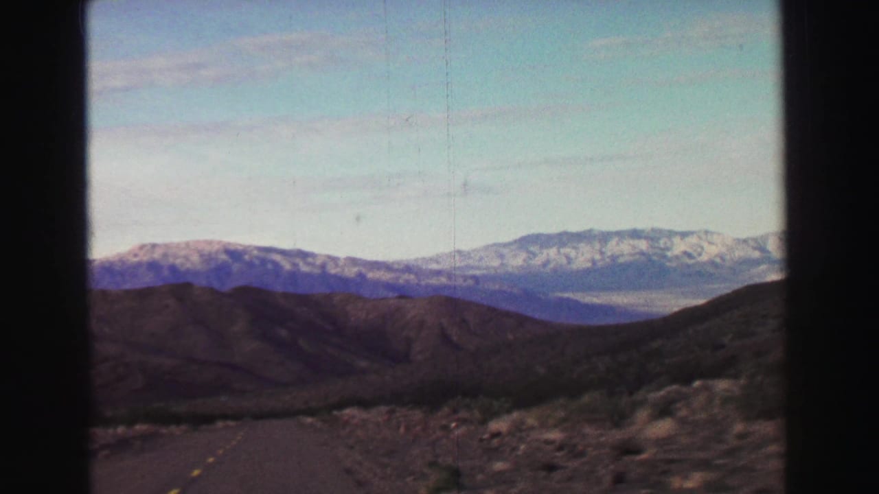 Two-lane mountain road leading toward distant snow-capped peaks under a pale sky.