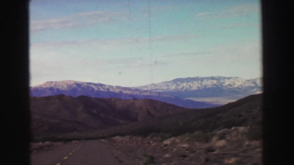 1969 Lake Tahoe-area 8mm view from a moving car, with a dark road foreground and snow-capped mountains beyond arid hills.