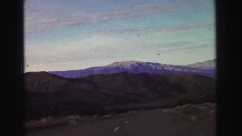 Lake Tahoe-area 1969 home-movie frame with a road dipping through hills toward a broad snow-capped ridge.