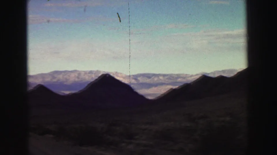 1969 8mm road-trip view near Lake Tahoe, with dark foreground peaks, pale blue sky, and distant mountain ridges.