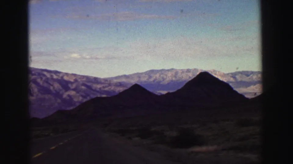 1969 Lake Tahoe-area road view from a moving vehicle, with yellow center lines, dark peaks, scrubland, and distant mountains.