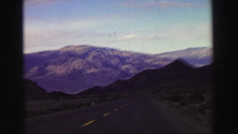 1969 8mm moving-car view near Lake Tahoe, showing a curved road, roadside scrub, dark peak, and layered mountain backdrop.