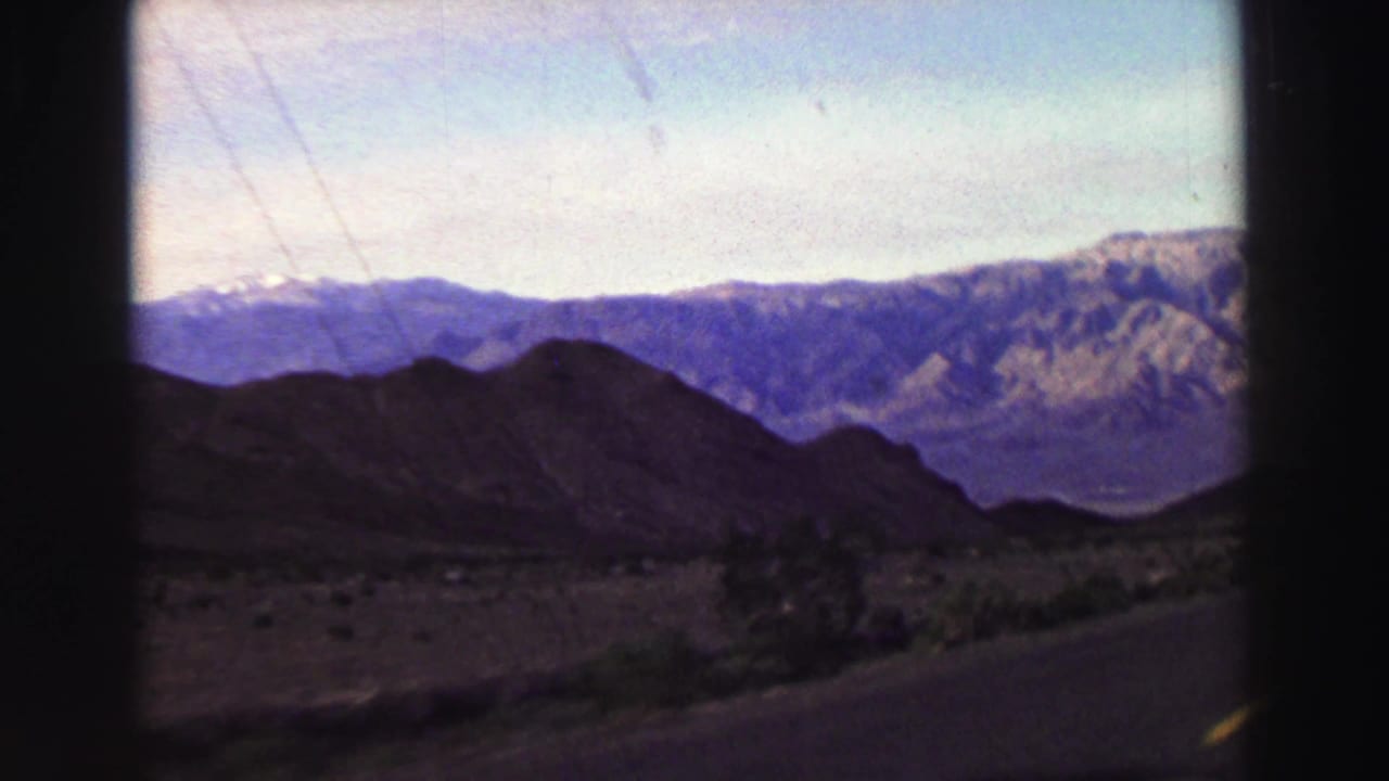 Paved road curving past dark hills with distant mountain ridges and pale sky.