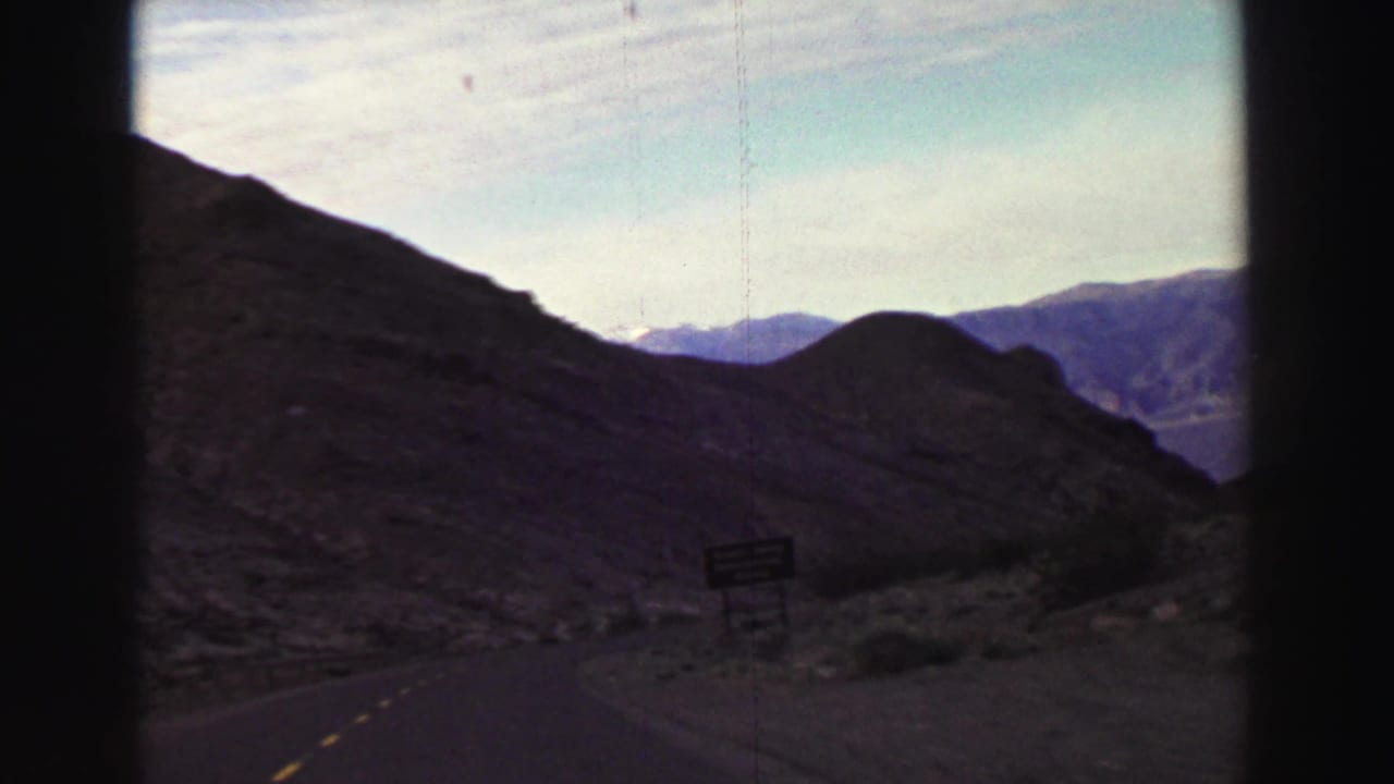 Mountain road curving past rocky hills with a small roadside sign and distant ridges.
