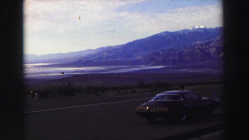 1969 Lake Tahoe roadside scene with a dark car in the foreground and mountains rising beyond the hazy lake.