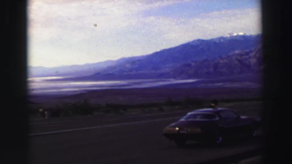 1969 Lake Tahoe-area home-movie frame showing a dark coupe on a highway shoulder below distant mountains.