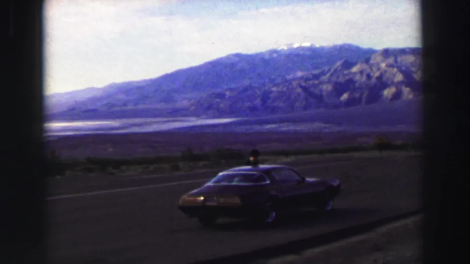 1969 Lake Tahoe-area frame with a parked dark coupe, highway pavement, and broad mountain landscape viewed through dark frame edges.