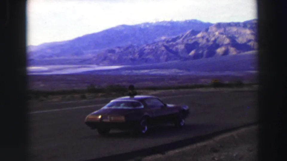 1969 Lake Tahoe-area home-movie frame showing the parked coupe and a person near it beside a broad mountain view.