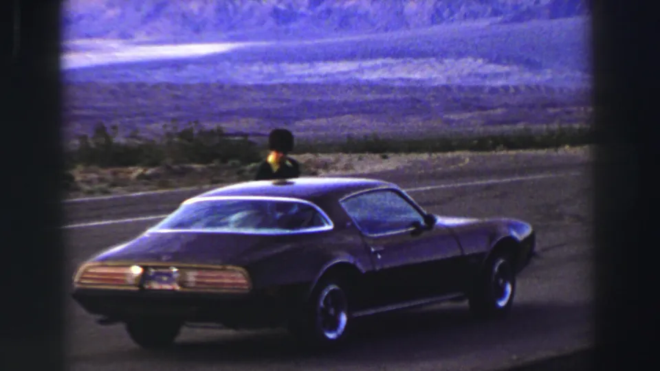 Closer 1969 Lake Tahoe-area frame of a dark parked coupe with a person visible near the roofline and mountains beyond.