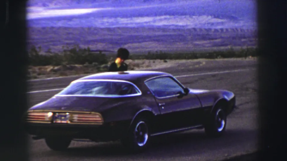 1969 Lake Tahoe home-movie frame showing a dark Pontiac Firebird beside a road as a person leans near the windshield area.