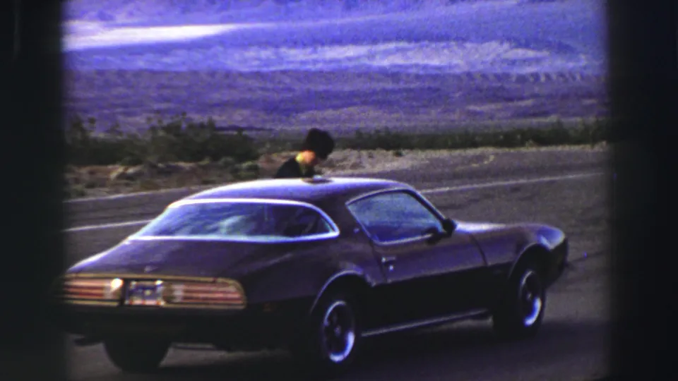 The dark Pontiac Firebird remains parked by the road while a person stands near the front window in the Lake Tahoe footage.