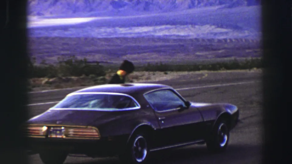 A grainy 8mm frame shows the Firebird from the rear side as the person leans toward the windshield or roofline.