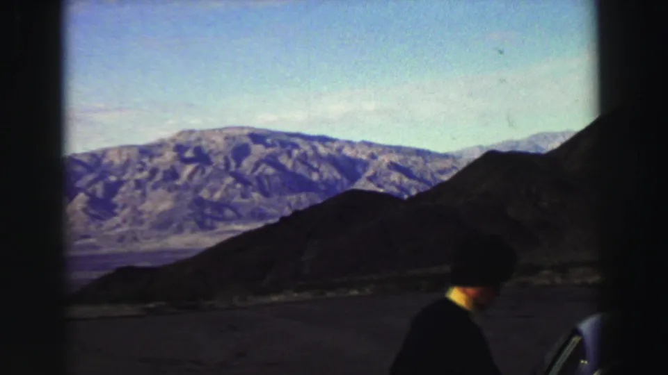 A 1969 home-movie frame shows rugged mountains and a roadside view, with a partially visible person near a vehicle at the lower edge.