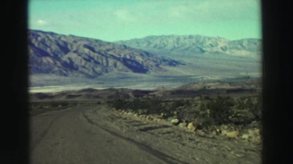 A winding road crosses dry scrubland toward rocky mountains in a 1969 Lake Tahoe-area 8mm travel view.