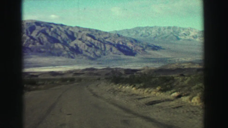 1969 8mm view of a road running through sparse California scrub toward a broad mountain range under a pale sky.