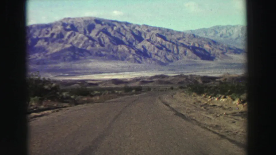 Moving 8mm frame from the California travel clip showing a road curving through dry brush toward rocky mountains.