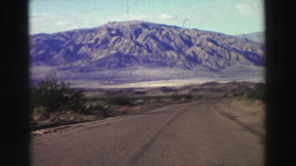 1969 home-movie frame showing a straight road, scrubby roadside, and a large mountain range beyond an open valley.