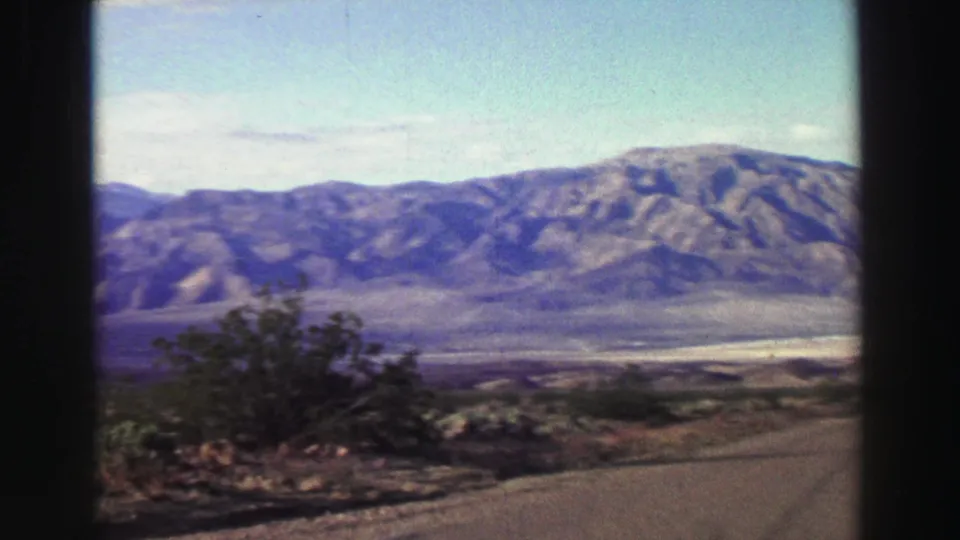 Late-1960s 8mm travel frame with a roadside shrub in the foreground and mountains across the open California landscape.