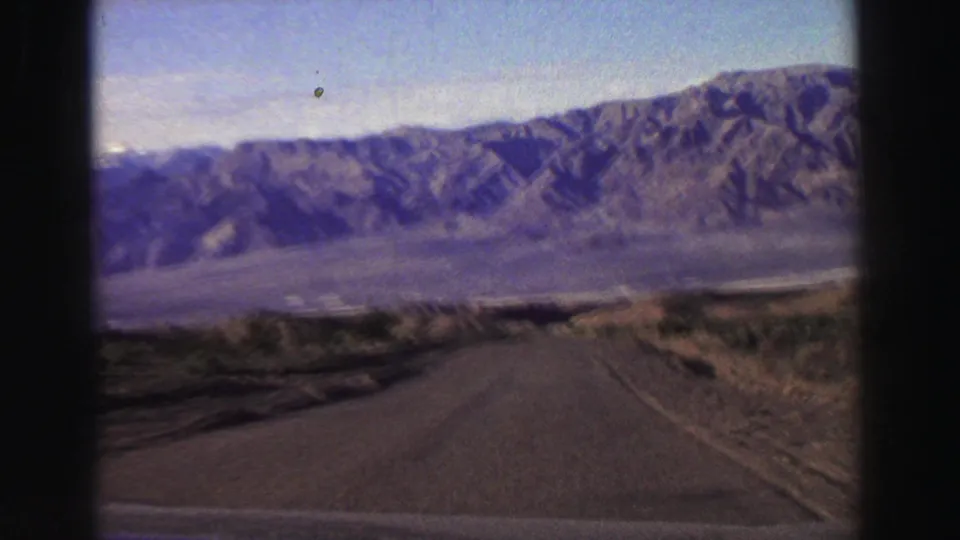 1969 Lake Tahoe-area home-movie frame showing a paved road receding toward rugged mountains and dry roadside brush.