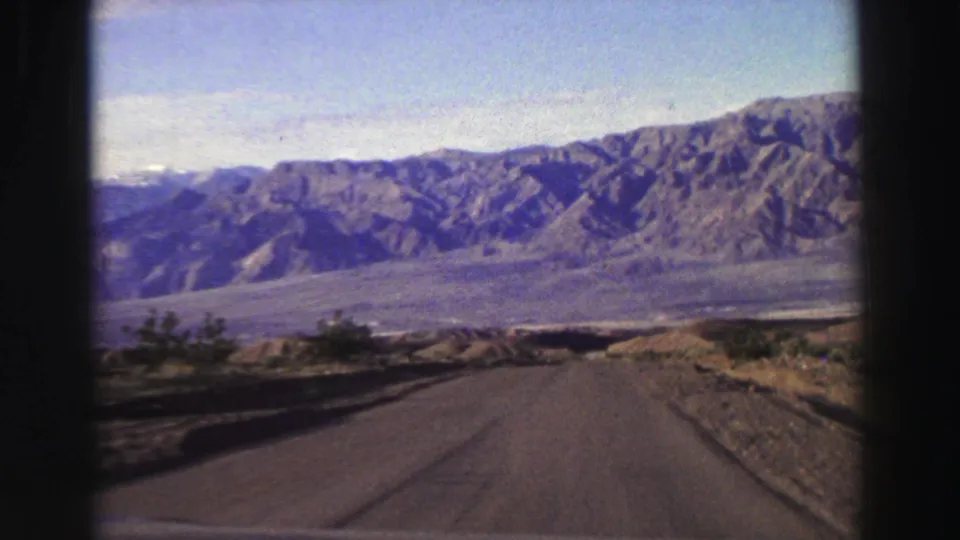 1969 Lake Tahoe-area road scene with a paved lane, dry shoulders, shrubs, valley floor, and rugged mountain ridge ahead.