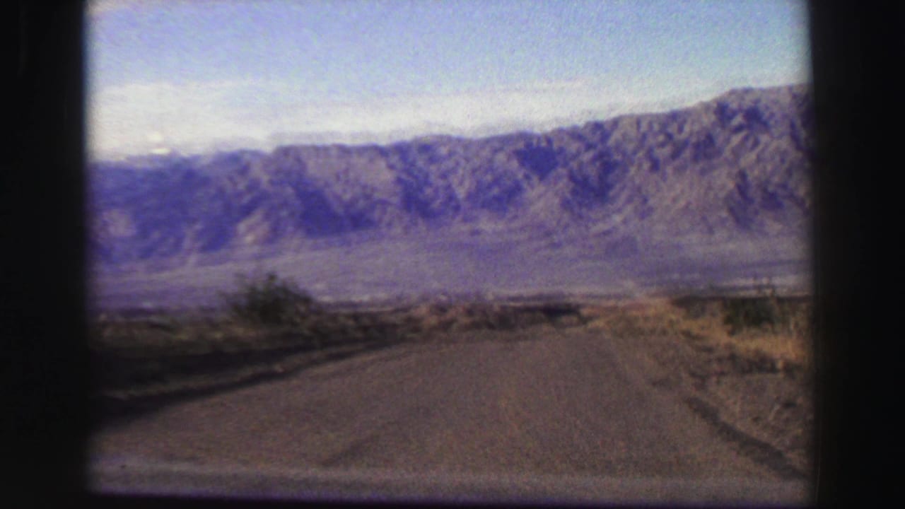 Paved road seen through a vehicle window with distant purple-brown mountains under a bright sky.