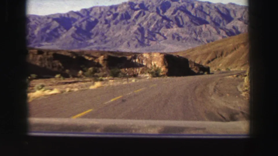 1969 Lake Tahoe-area frame of a vehicle approaching a curve through arid hills, with rocky roadside cuts and mountain ridges in the background.