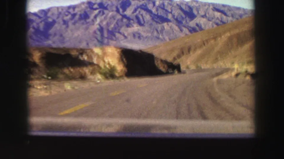 Home-movie frame from a 1969 California mountain drive, showing the road bending past a rocky slope with purple-brown peaks behind it.