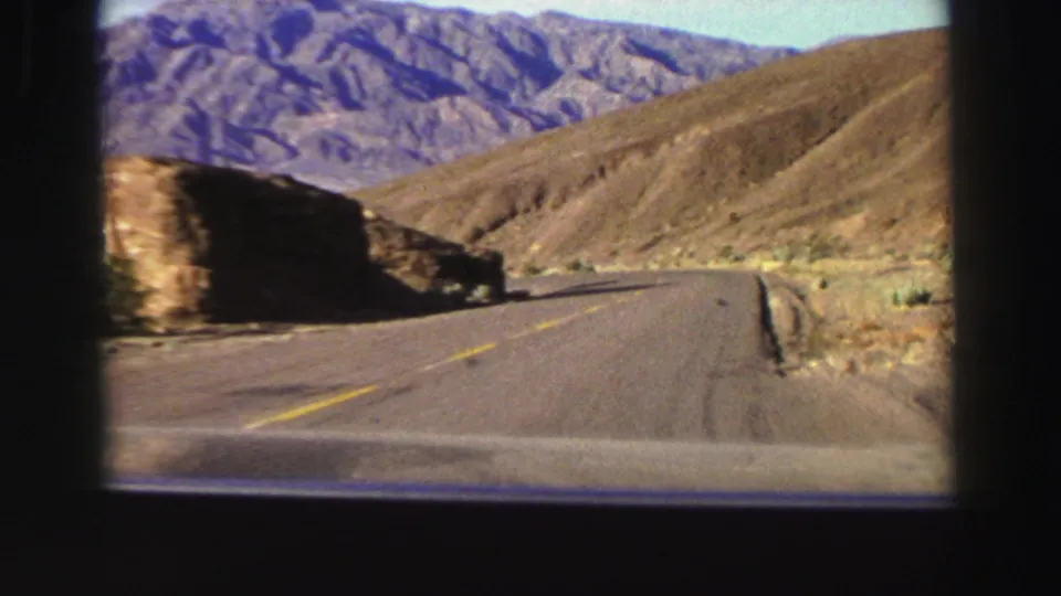 1969 Lake Tahoe-area road footage showing asphalt, a roadside edge, dry slopes, and purple-hued mountain ridges from a moving vehicle.