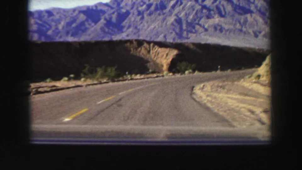 Driver’s-eye 8mm view of a curving Lake Tahoe-area mountain road with sparse roadside vegetation and rugged hills beyond.