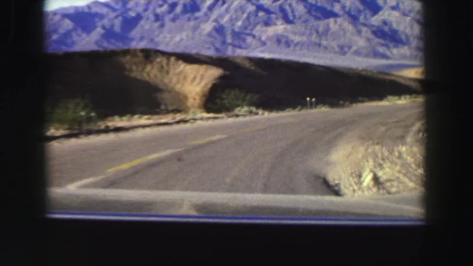 Forward road-trip view in the Lake Tahoe area, showing a paved bend, dry shoulder, sparse shrubs, and mountain slopes in 8mm color.