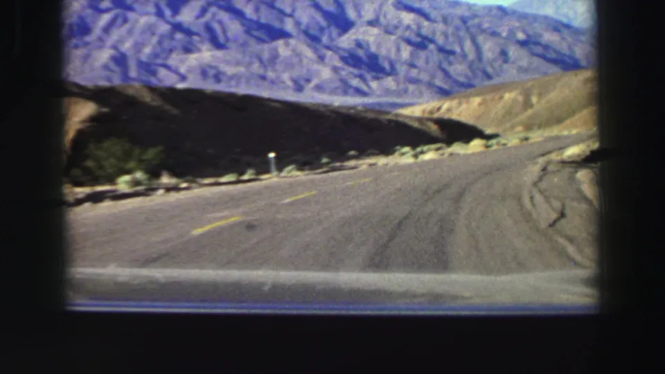 1969 8mm vehicle perspective on a winding Lake Tahoe-area road, with tan hillsides, sparse vegetation, and distant rugged mountains.